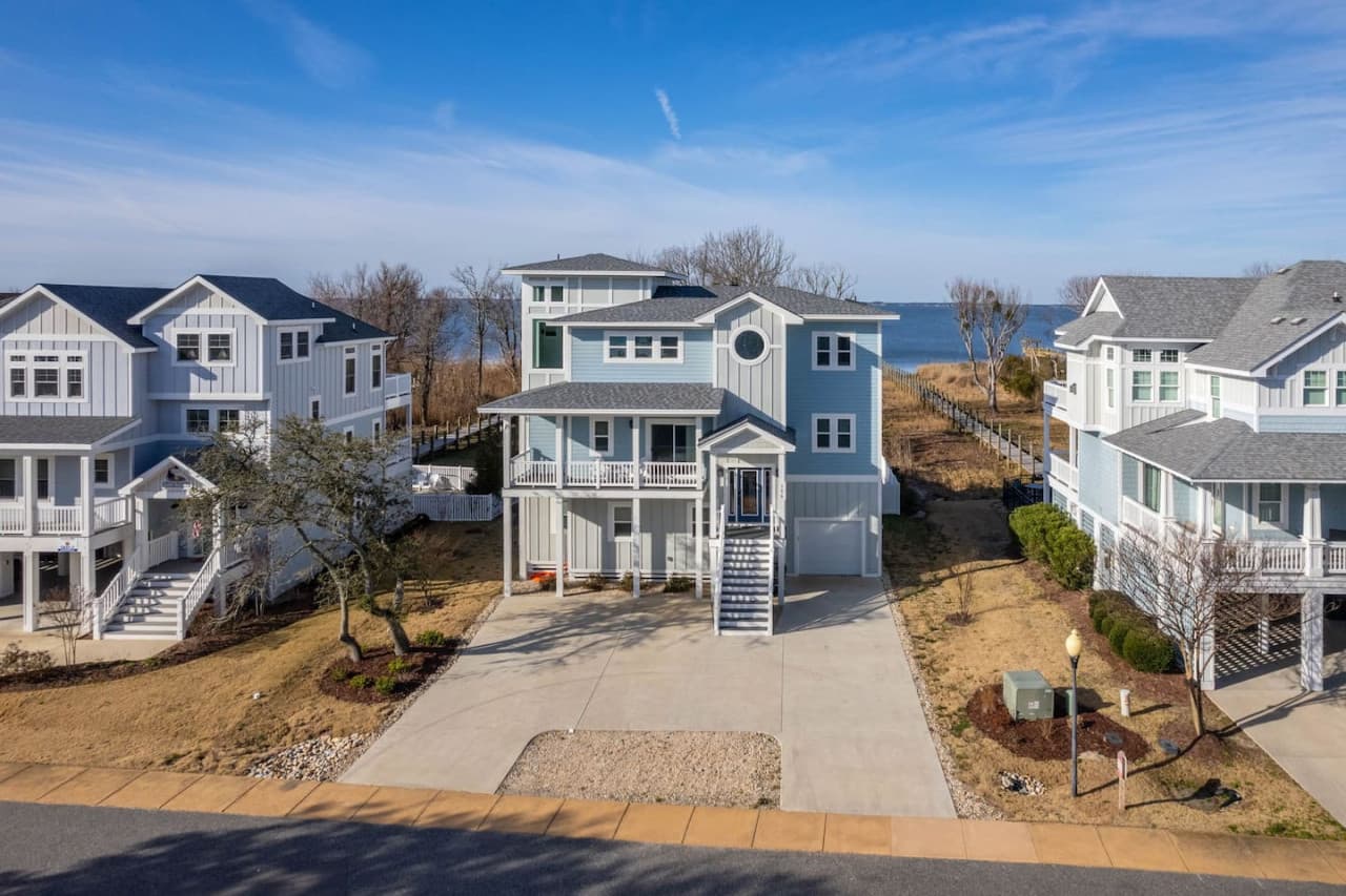 Street view of the blue-sided home w/ covered porches, private drive & the sound behind.