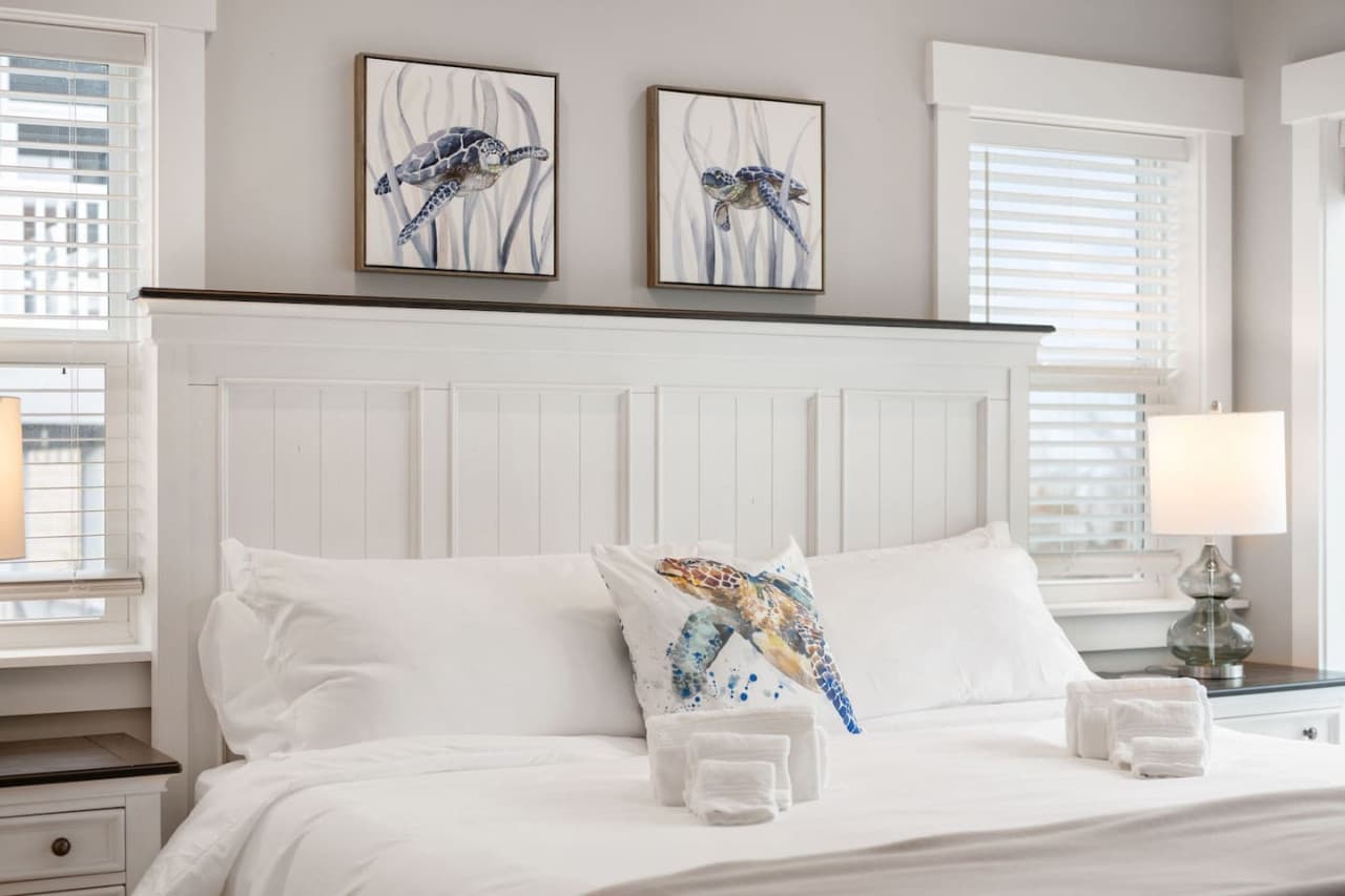 Bedroom w/ white panelled headboard, coastal prints & fresh towels set out on the bed.