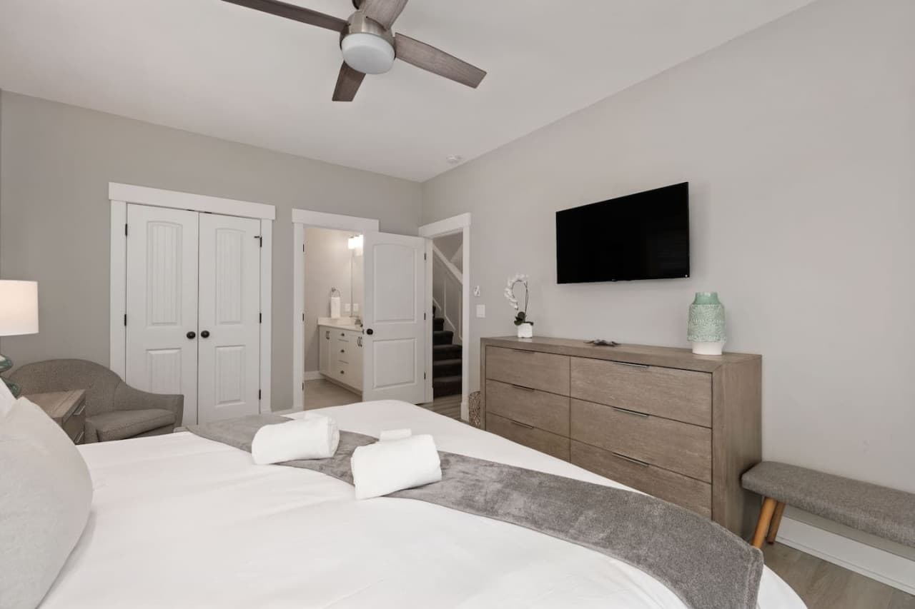 Bedroom w/ mounted TV, wide wood dresser & a padded bench at the foot of the bed.