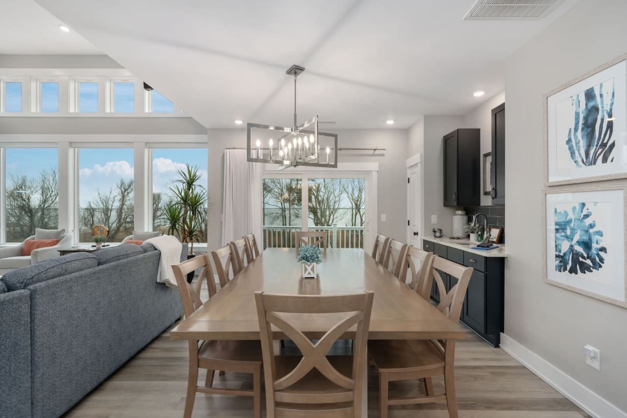 Living & dining area w/ blue sofa, long farmhouse table & coastal art for group meals.