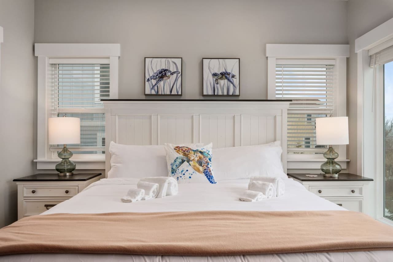 Bedroom w/ paired glass lamps, framed coastal prints & a blush throw across the bed.