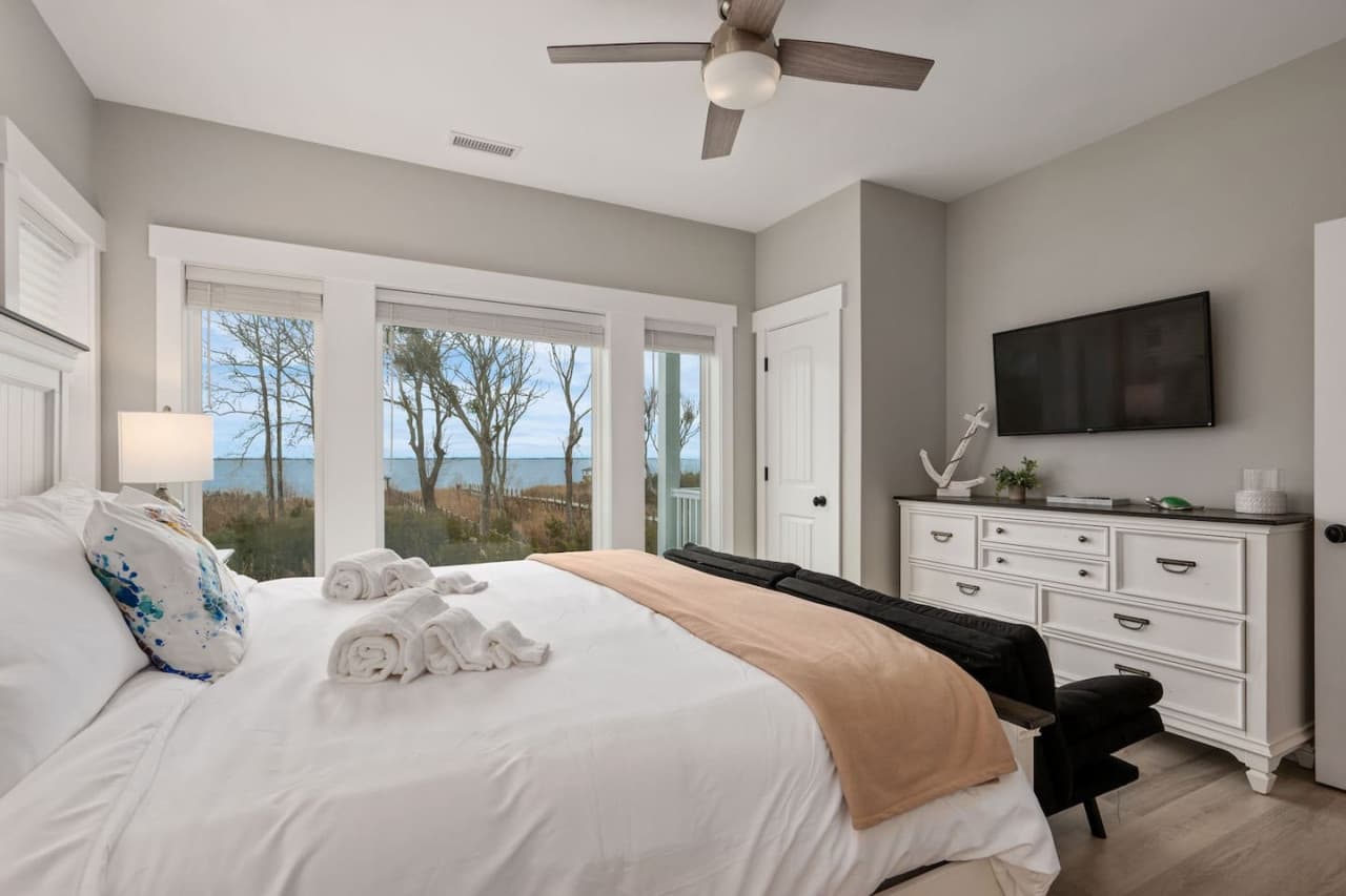 Bedroom w/ a wall of windows over the sound, mounted TV & white dresser.