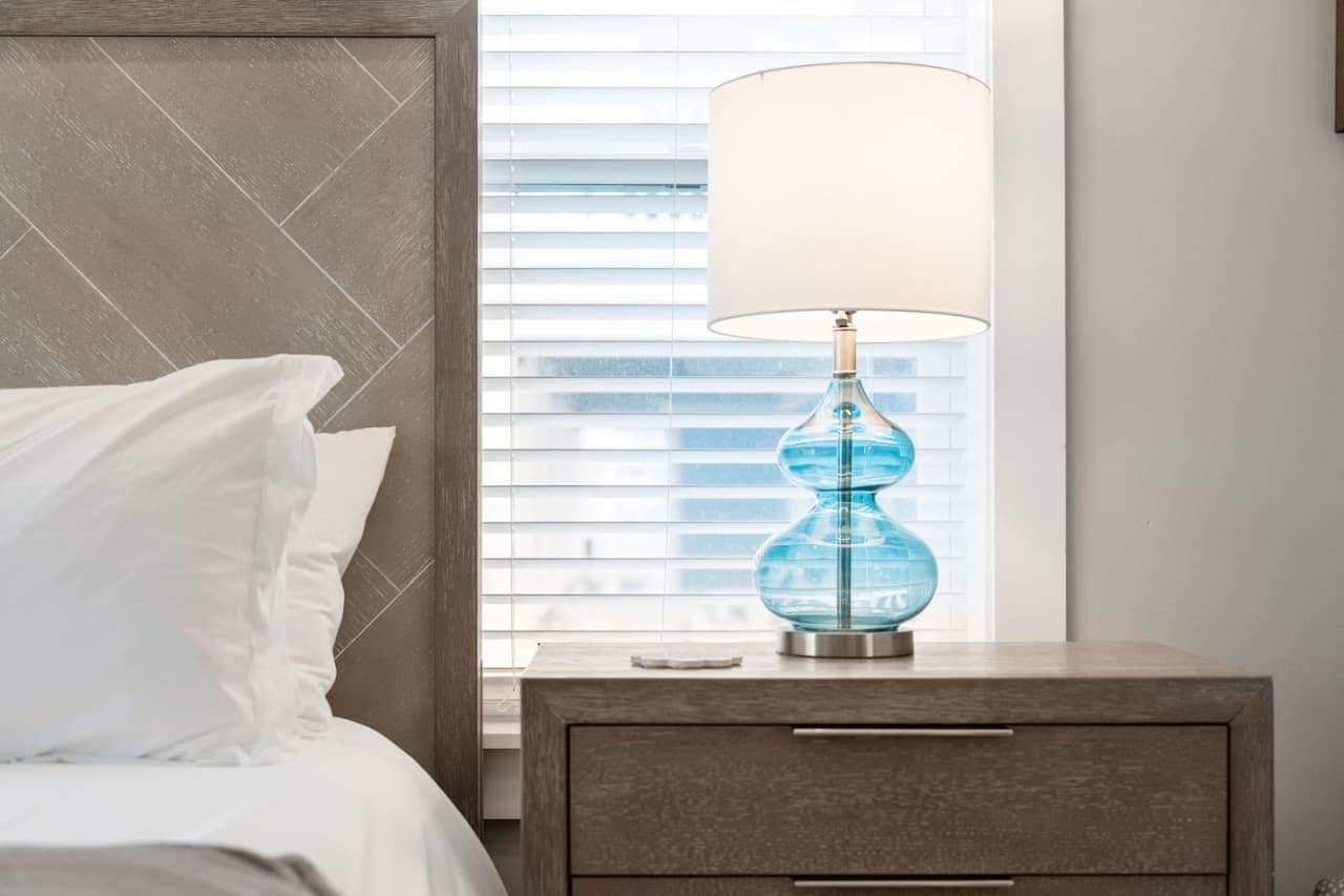 Bedside detail w/ blue glass lamp, wood nightstand & soft light through the blinds.