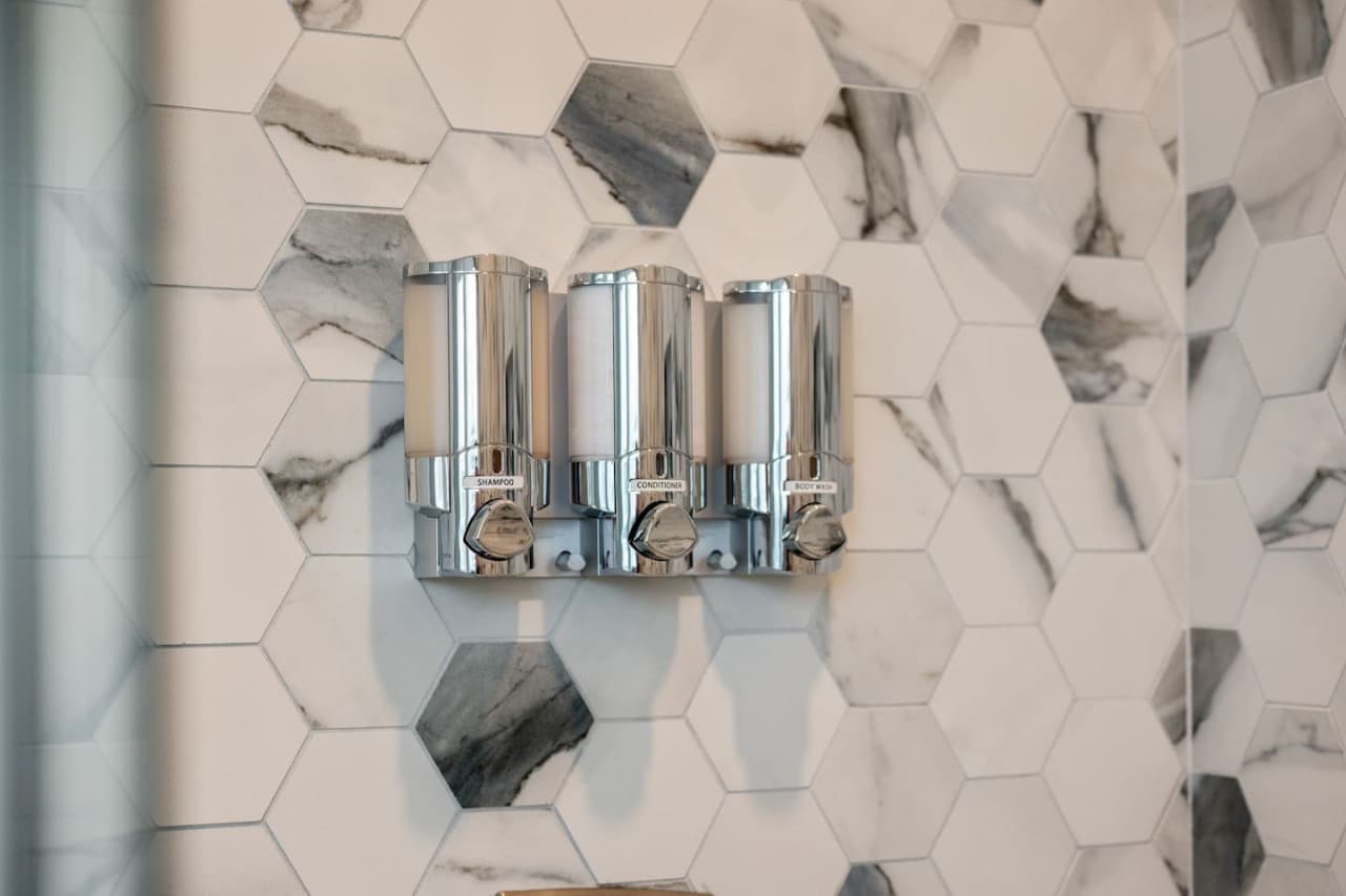 Shower detail w/ hex marble-look tile & wall-mounted shampoo, conditioner & body wash.