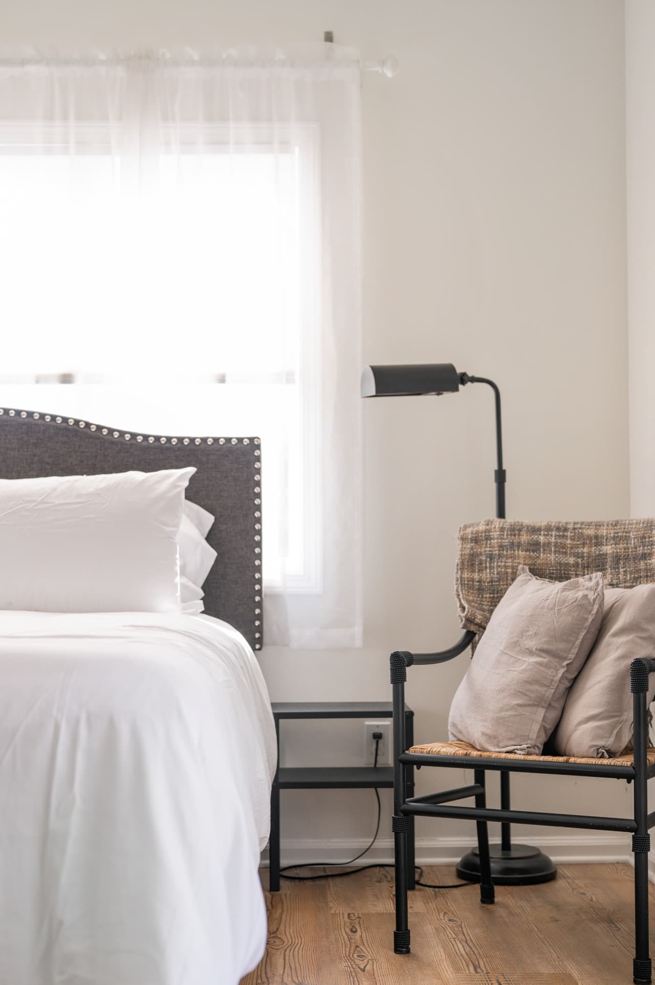 Cozy bedroom corner w/ layered pillows, reading lamp, & soft natural light.