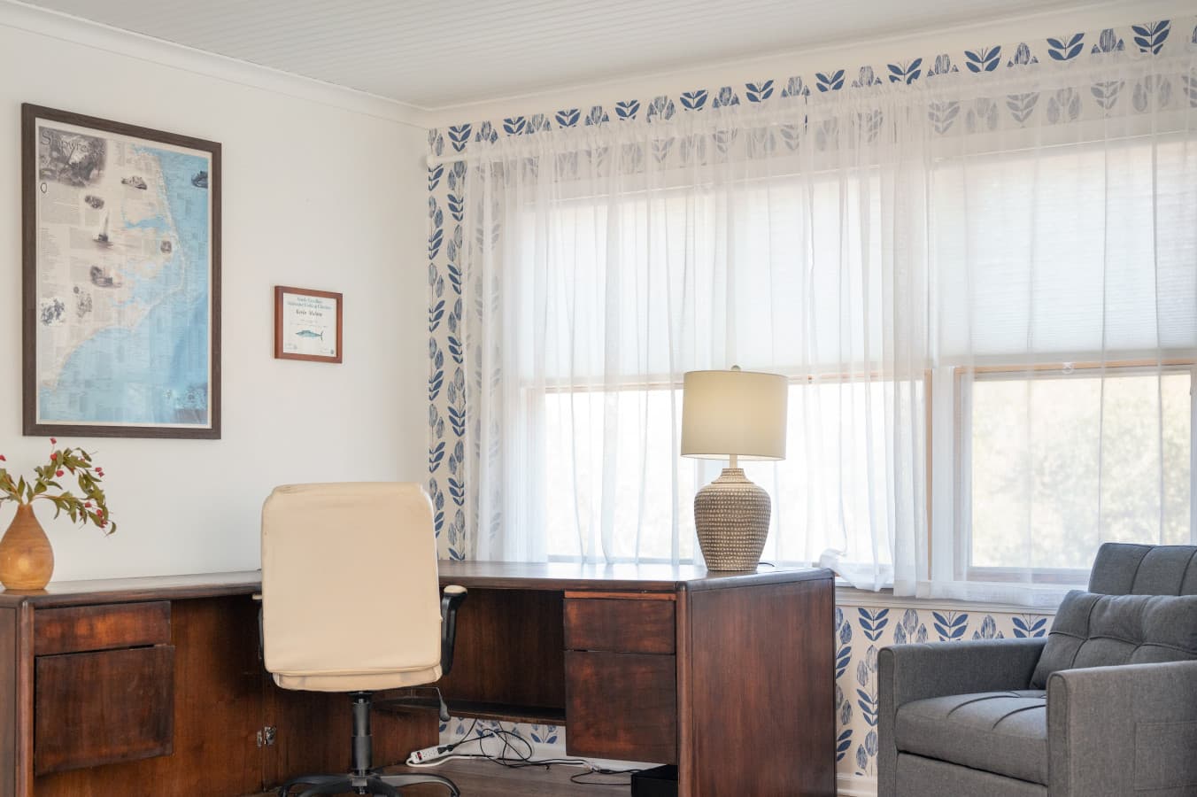 Desk nook w/ wood desk, task chair & lamp beneath bright windows for catching up on work.