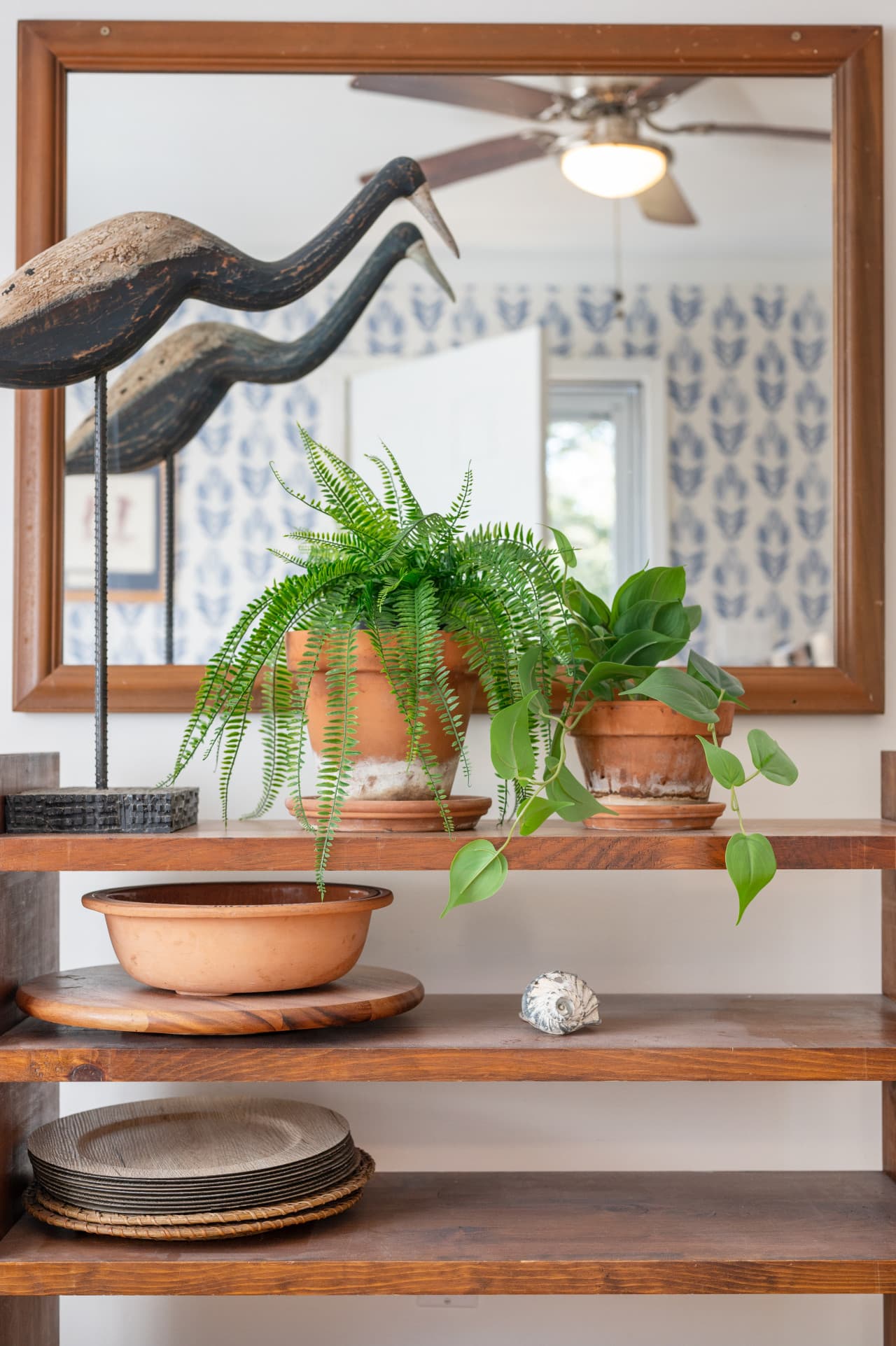 Coastal decor detail w/ potted ferns, a pelican sculpture & mirror above an open wood shelf.
