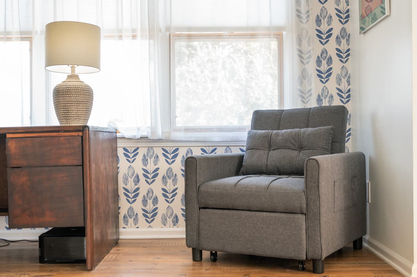 Reading corner w/ upholstered armchair, wood dresser & lamp beside a bright window.