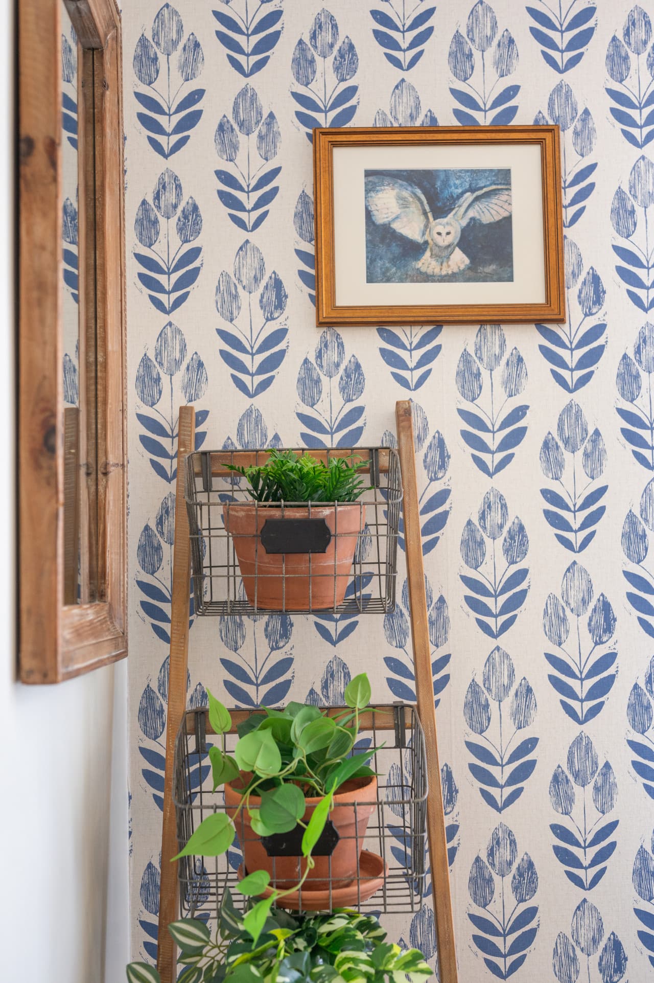 Decor detail w/ blue botanical wallpaper, a framed bird print & a ladder shelf of plants.