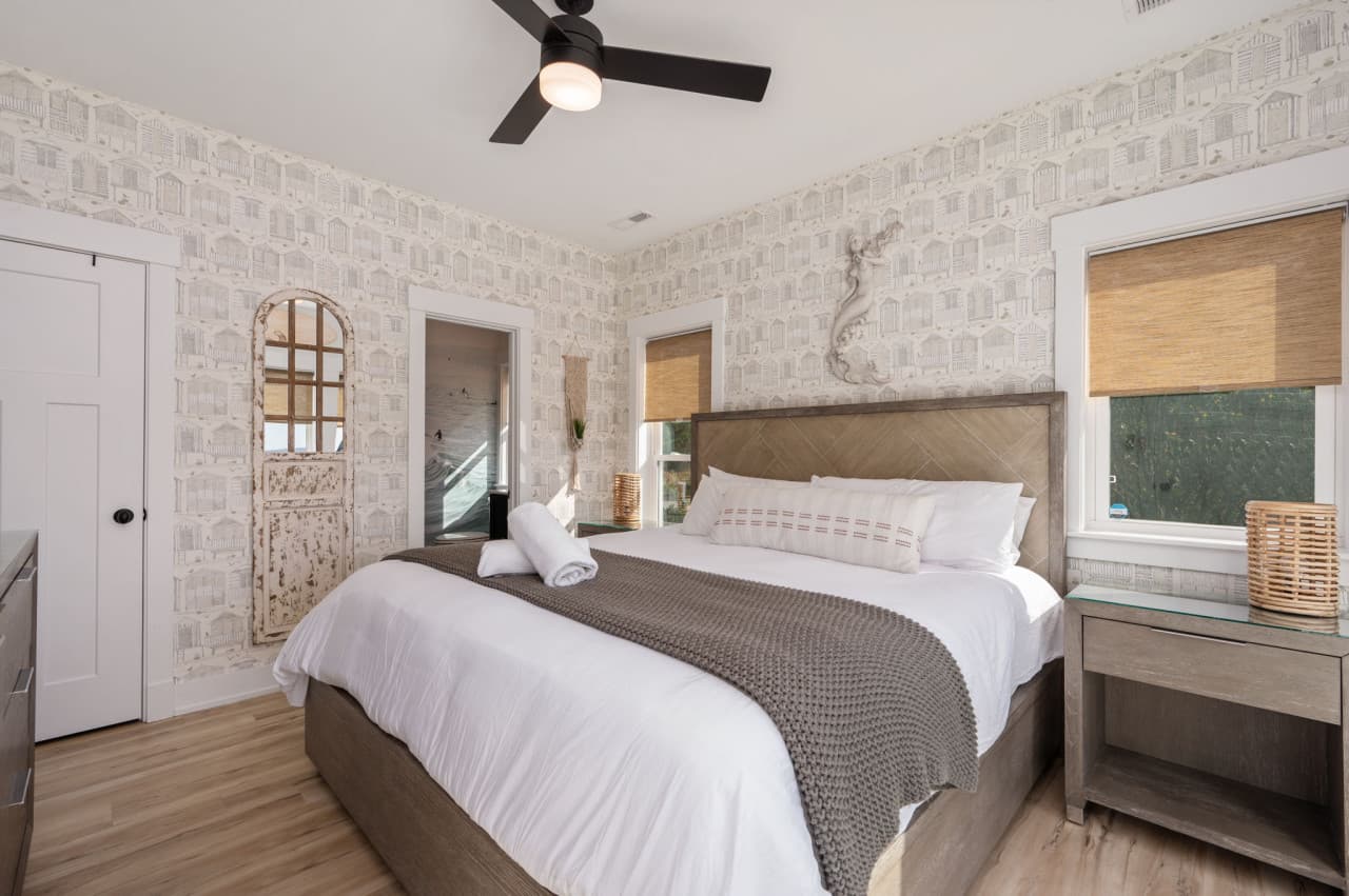 Bedroom w/ light wallpaper, woven window shades & a bench at the foot of the bed.