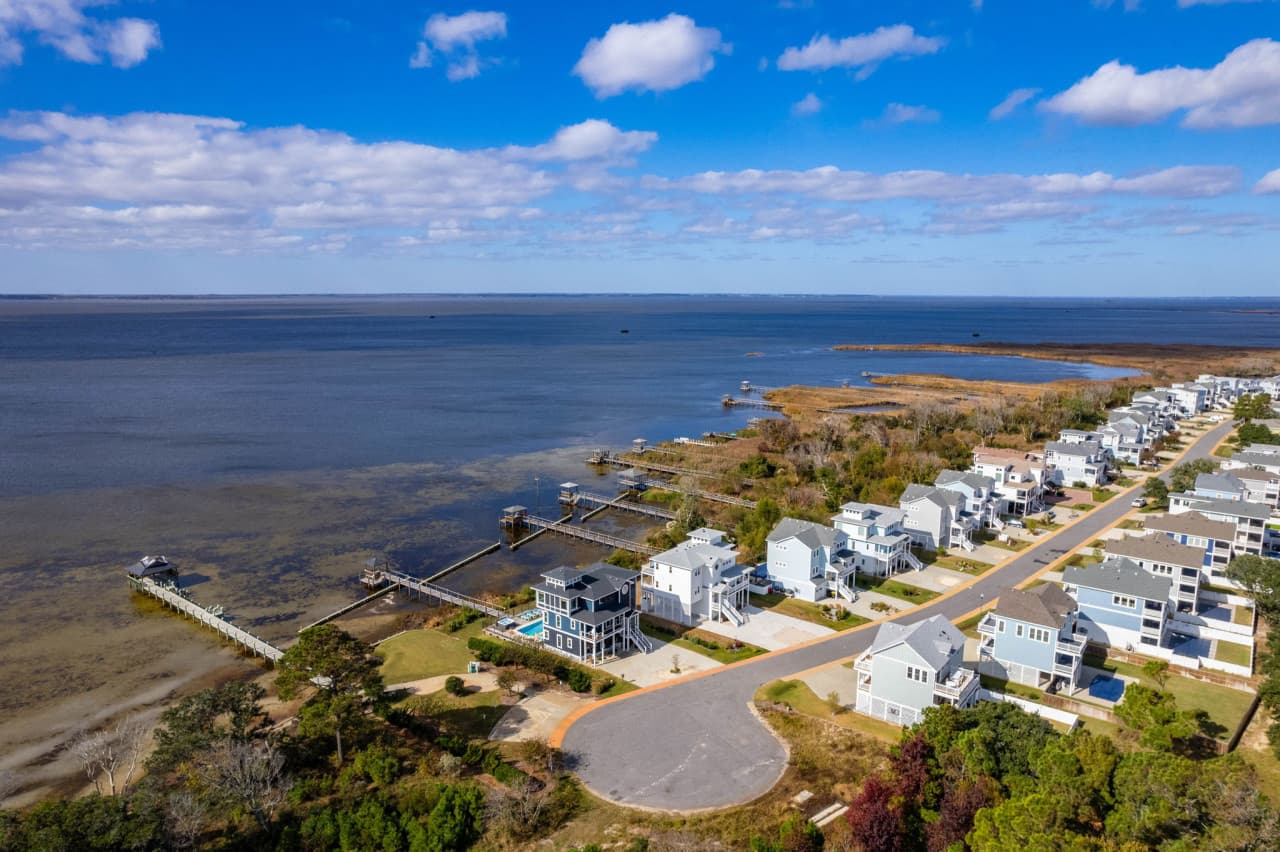 Aerial over the sound-side street w/ marsh, piers & open water just beyond.
