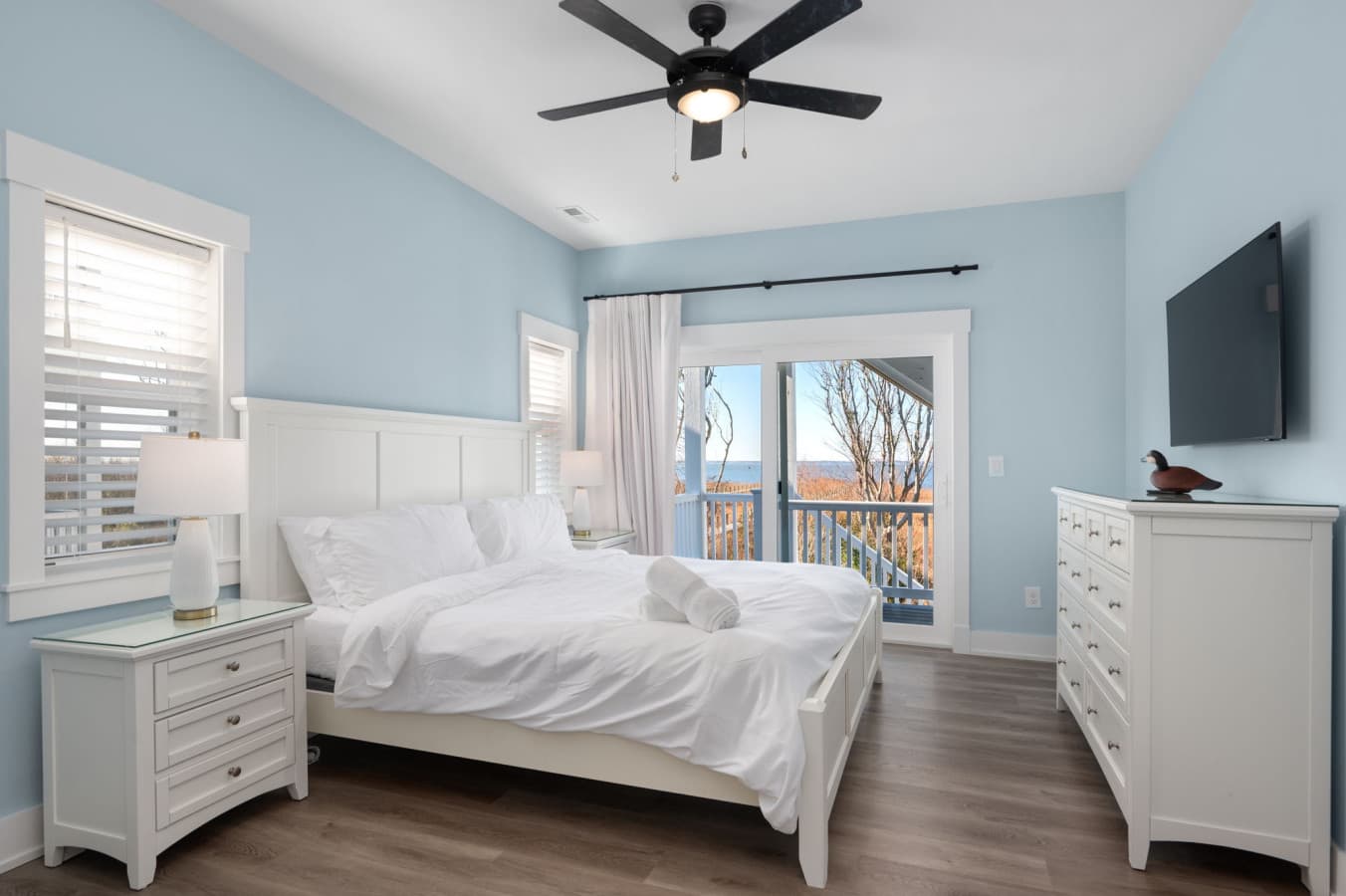 Bedroom w/ pale blue walls, wall TV & sliders opening onto a deck with water views