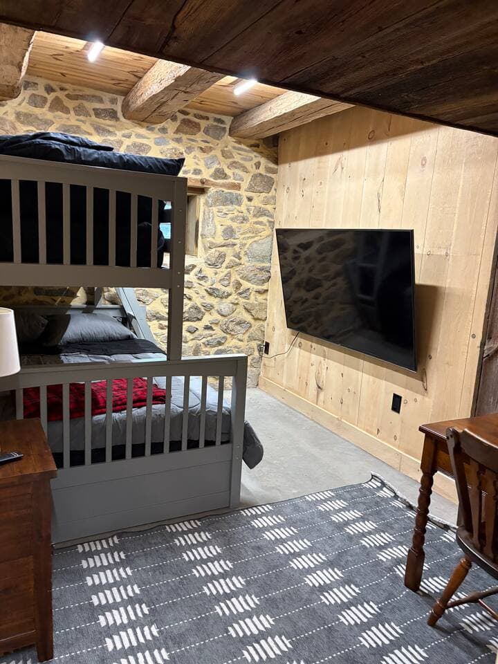 A spacious farmhouse-inspired bedroom featuring a sturdy bunk bed with one full bed and two single beds. Warm rustic décor and natural wood accents create a cozy barn-style atmosphere. The room also includes a large screen TV.