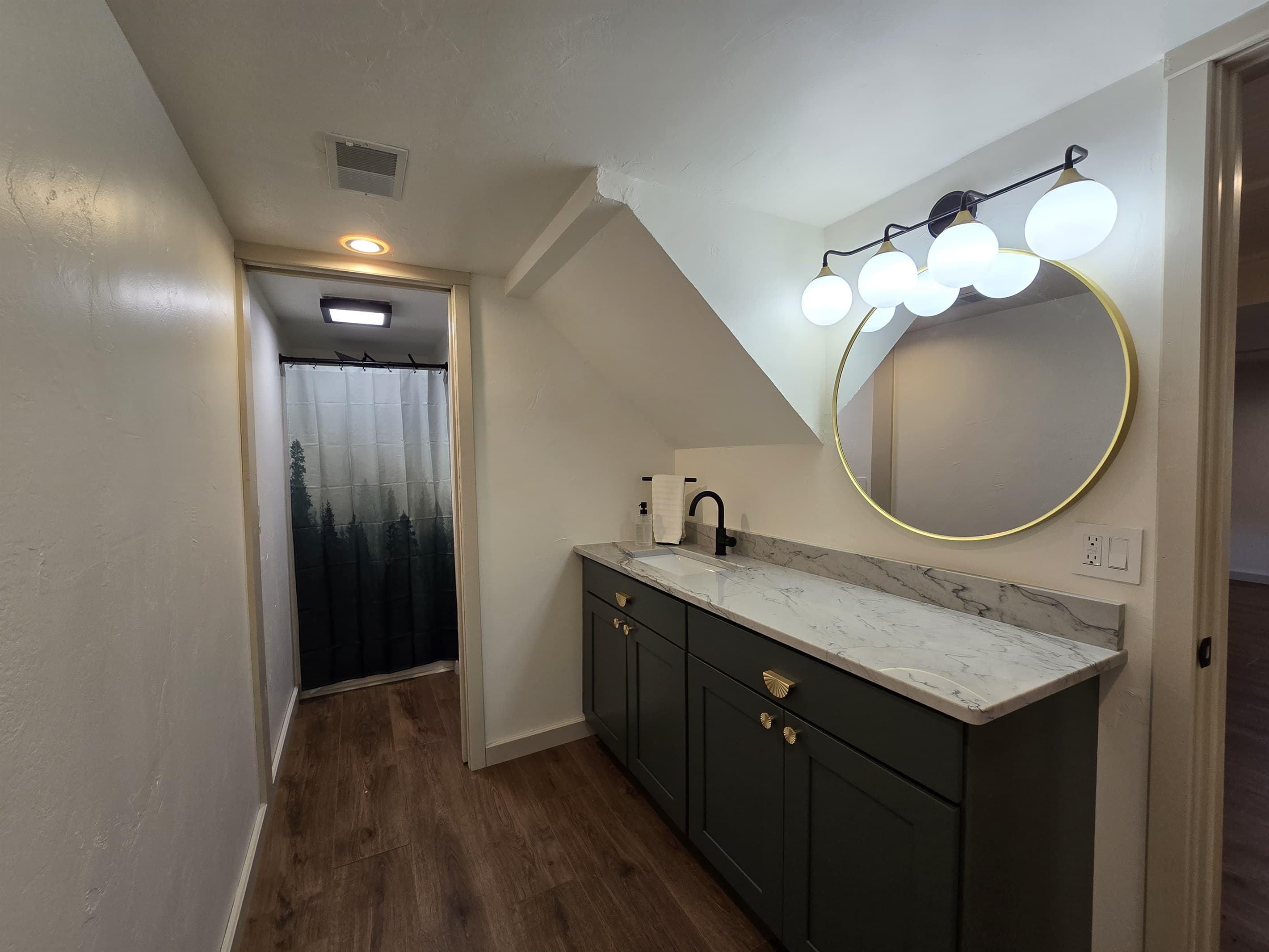 Spacious lake house bathroom with a stylish marble countertop and modern fixtures. The walk-in shower is highlighted by a forest-themed curtain. Black cabinetry with gold accents adds a touch of elegance. A large, round mirror with vanity lights enhances t