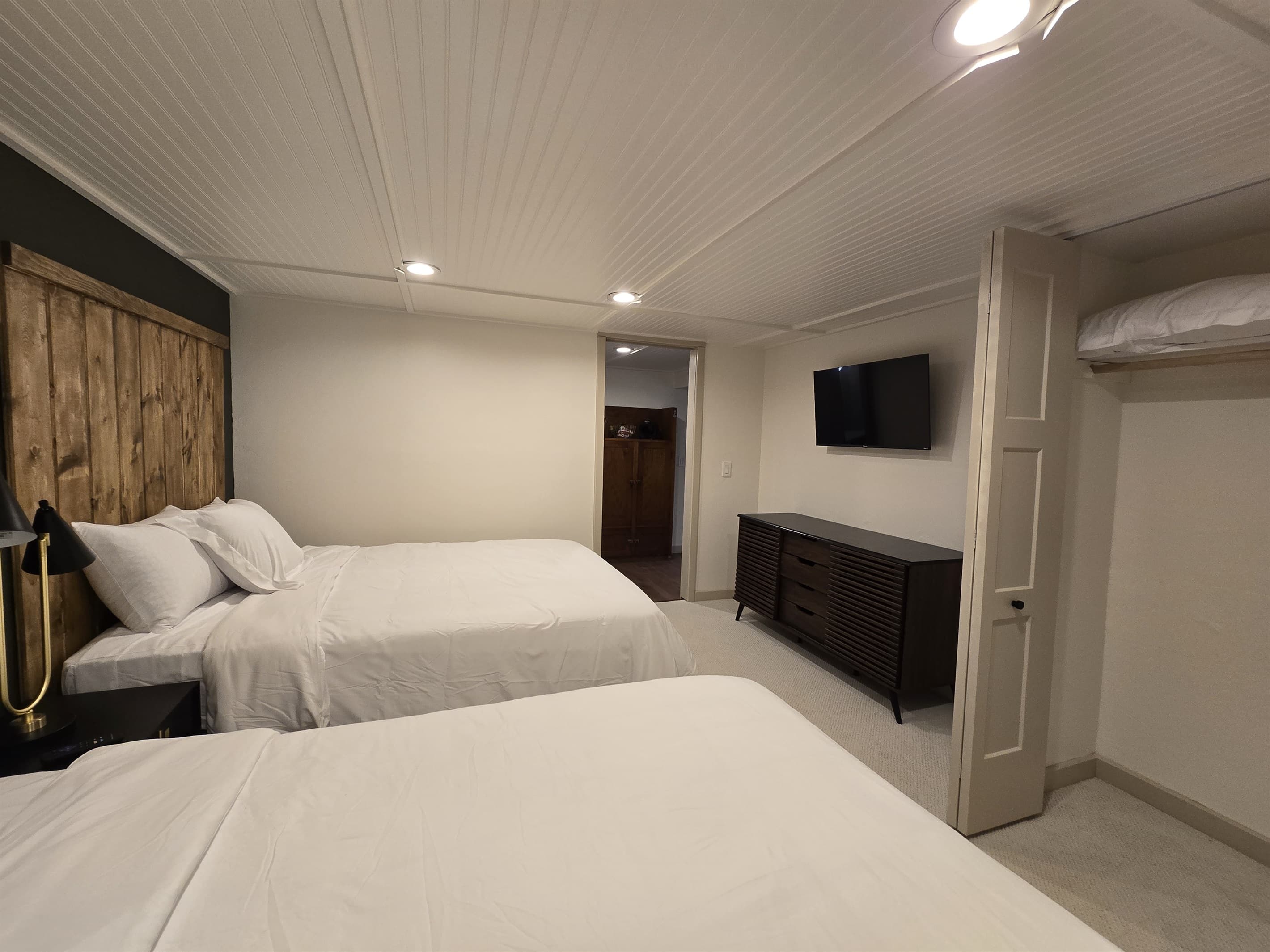 Comfortable lake house bedroom with two queen-sized beds, ideal for guests. Rustic wooden headboards add warmth, while the room is equipped with modern amenities like a flat-screen TV. The carpeted floor and recessed lighting create a cozy atmosphere.