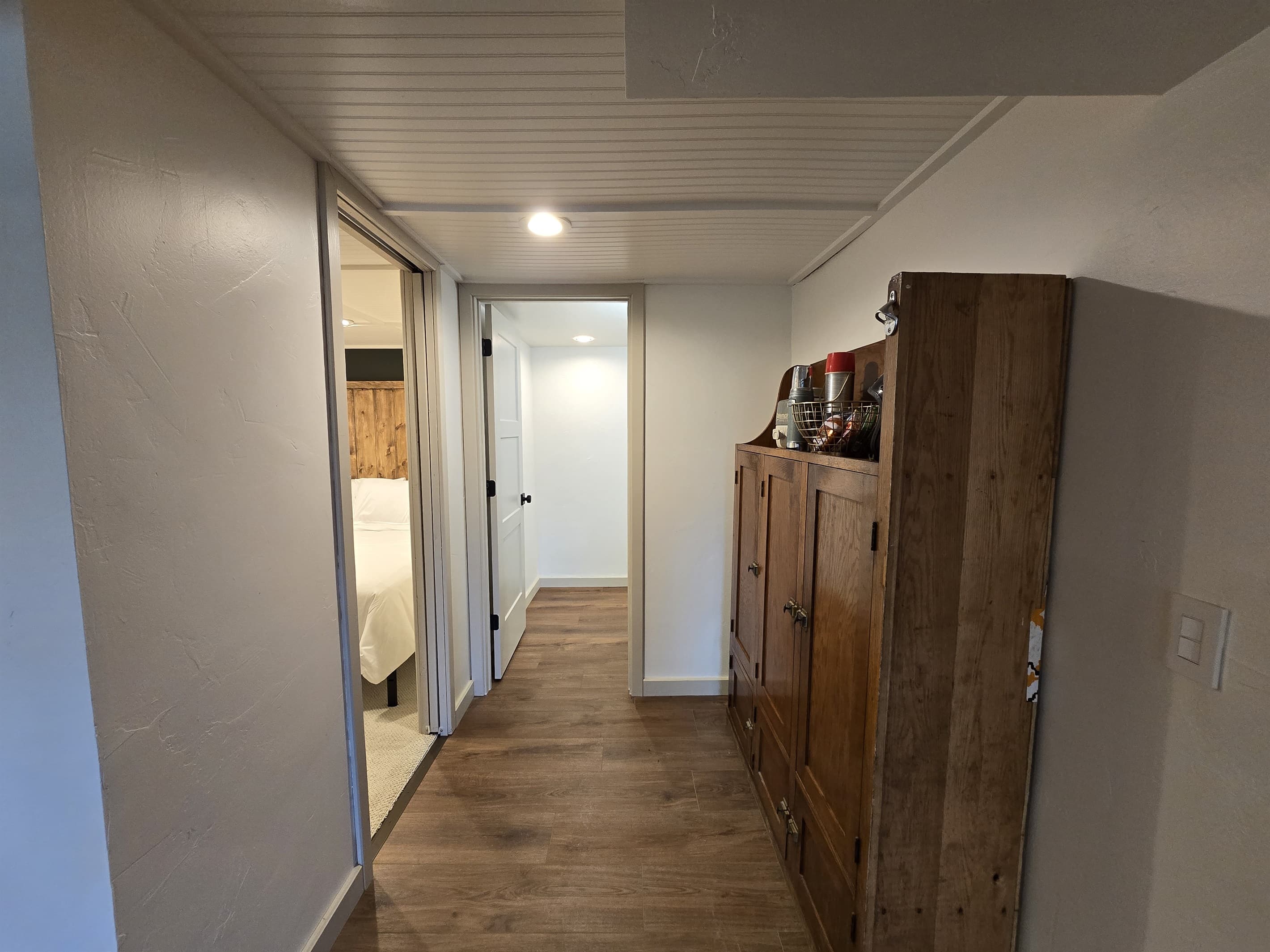 Cozy hallway in the lake house leading to a bedroom. Features a rustic wooden cabinet for storage, with decorative items on top. The hallway has wooden flooring and leads to a bedroom with visible natural light, providing a welcoming feel.