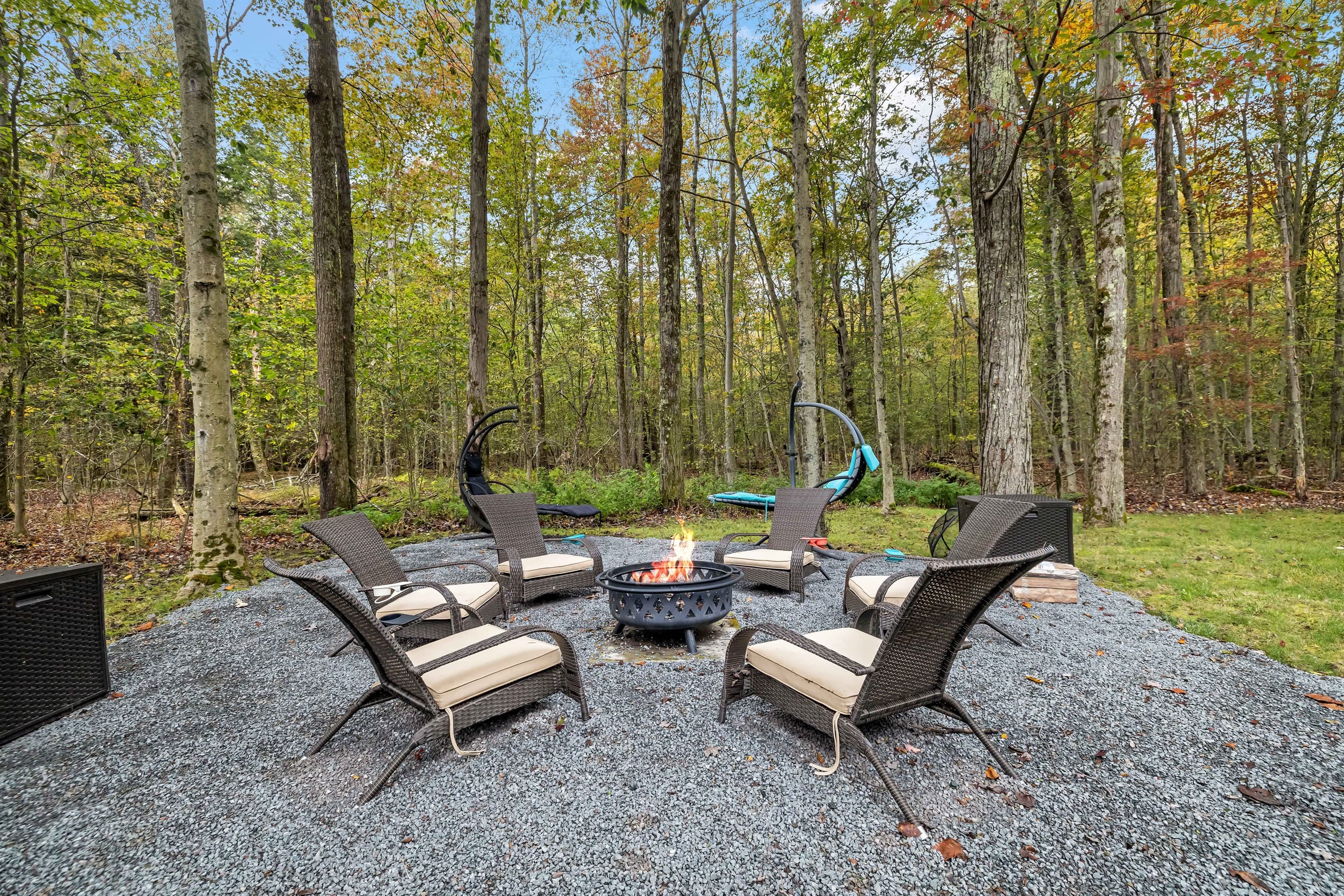 Pocono mountains rental | Relax in the spacious backyard featuring a firepit, chairs, and loungers, all with a serene forest view.