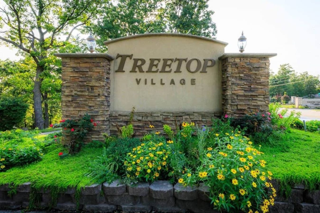 This inviting entrance to Treetop Village welcomes you with a beautifully landscaped monument sign, featuring charming stone pillars and lush greenery. The vibrant flowers and well-maintained surroundings immediately convey a sense of community and c