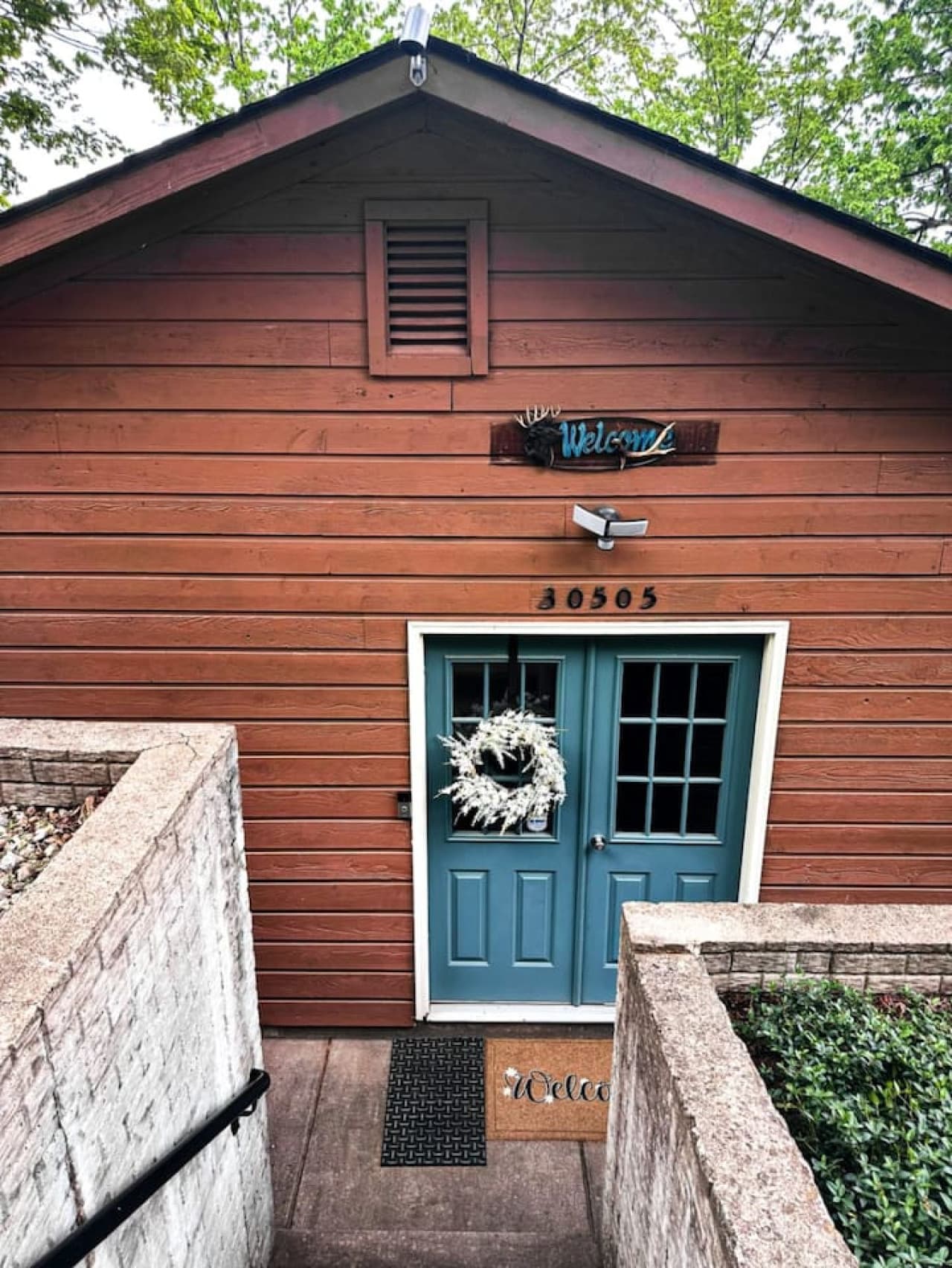 Step into this inviting lakeside cabin through a charming entryway featuring a cheerful blue double door, seasonal wreath, and rustic "Welcome" décor. The warm wood exterior and private walkway create a cozy, tucked-away feel that sets the tone for a