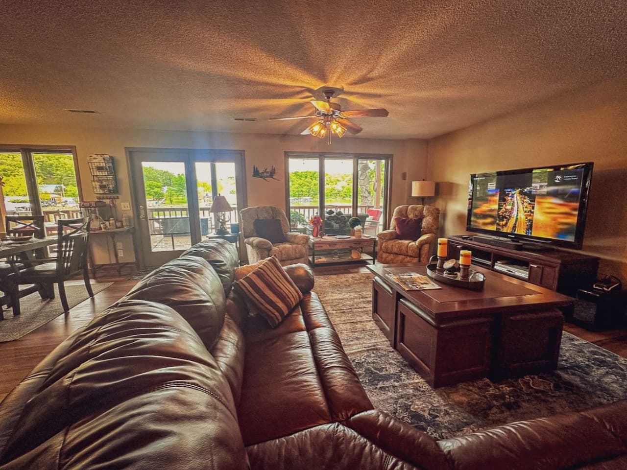 From this angle, the rustic living room opens up to reveal generous natural light streaming through large windows that frame scenic outdoor views. The blend of weathered wood furniture, soft throws, and vintage accents creates a welcoming space that
