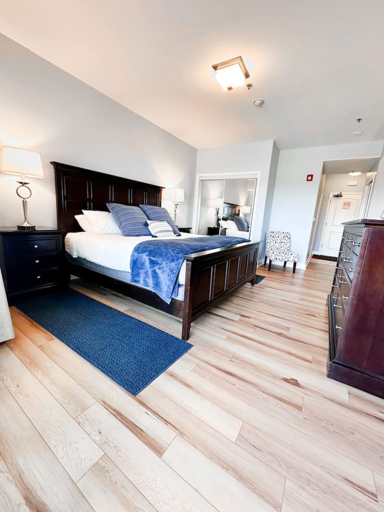 This dark mahogany bedroom features a large bed with blue bedding and a matching rug. A sliding mirrored closet and a dark wood dresser add to the room's elegance.