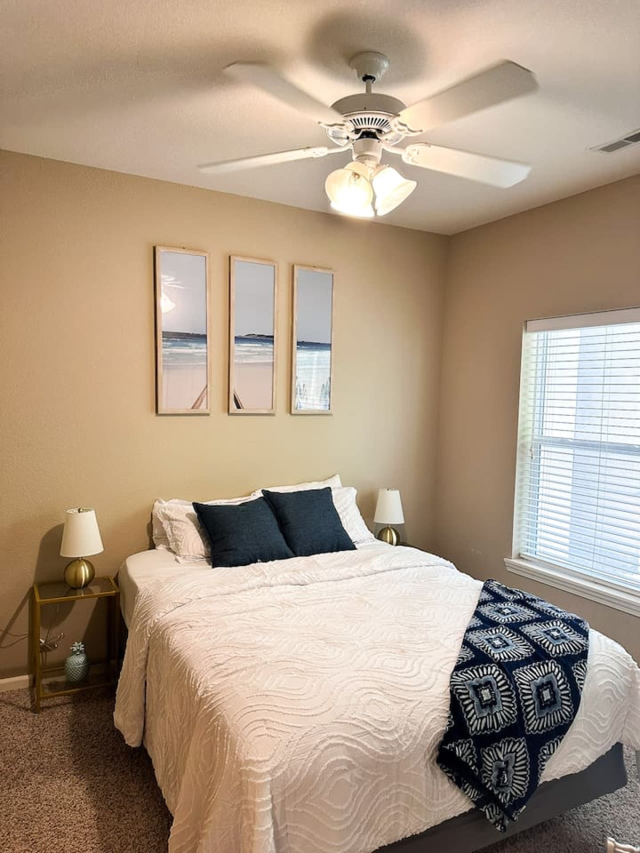 This inviting bedroom offers a comfortable queen-sized bed, complete with crisp white linens and decorative pillows for a restful night's sleep. The room is bathed in natural light from the large window, complemented by subtle wall art and a ceiling