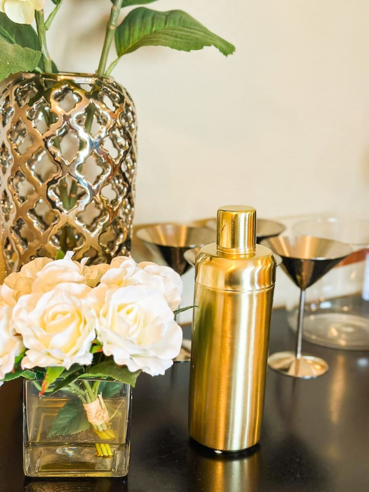 This elegant bar cart offers a sophisticated touch, featuring a selection of glasses and a sleek gold cocktail shaker, perfect for crafting your favorite drinks. Adorned with a beautiful vase of white flowers, it adds both functionality and style to
