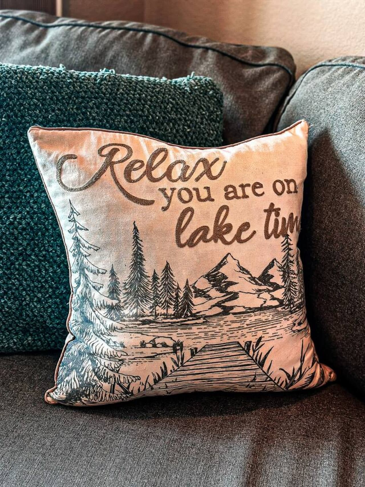This cozy throw pillow, featuring a charming lakeside design and a relaxing message, adds a touch of rustic comfort to the living space. Its inviting aesthetic perfectly complements the comfortable sofa, creating a warm and welcoming atmosphere for