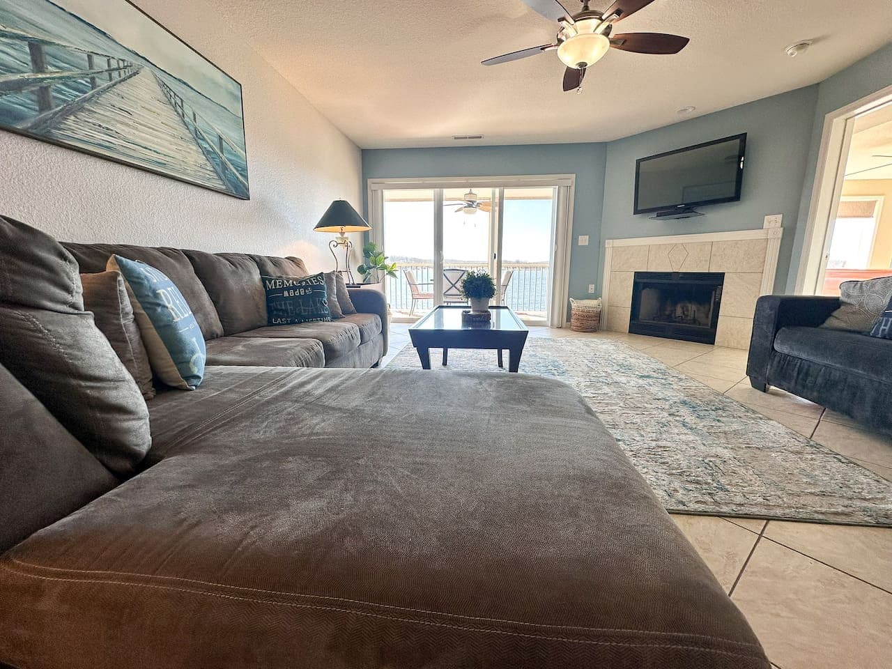 A spacious open-concept living area with a plush sectional sofa, a cozy fireplace, and a wall-mounted TV—all while offering stunning lakefront views through large sliding doors.