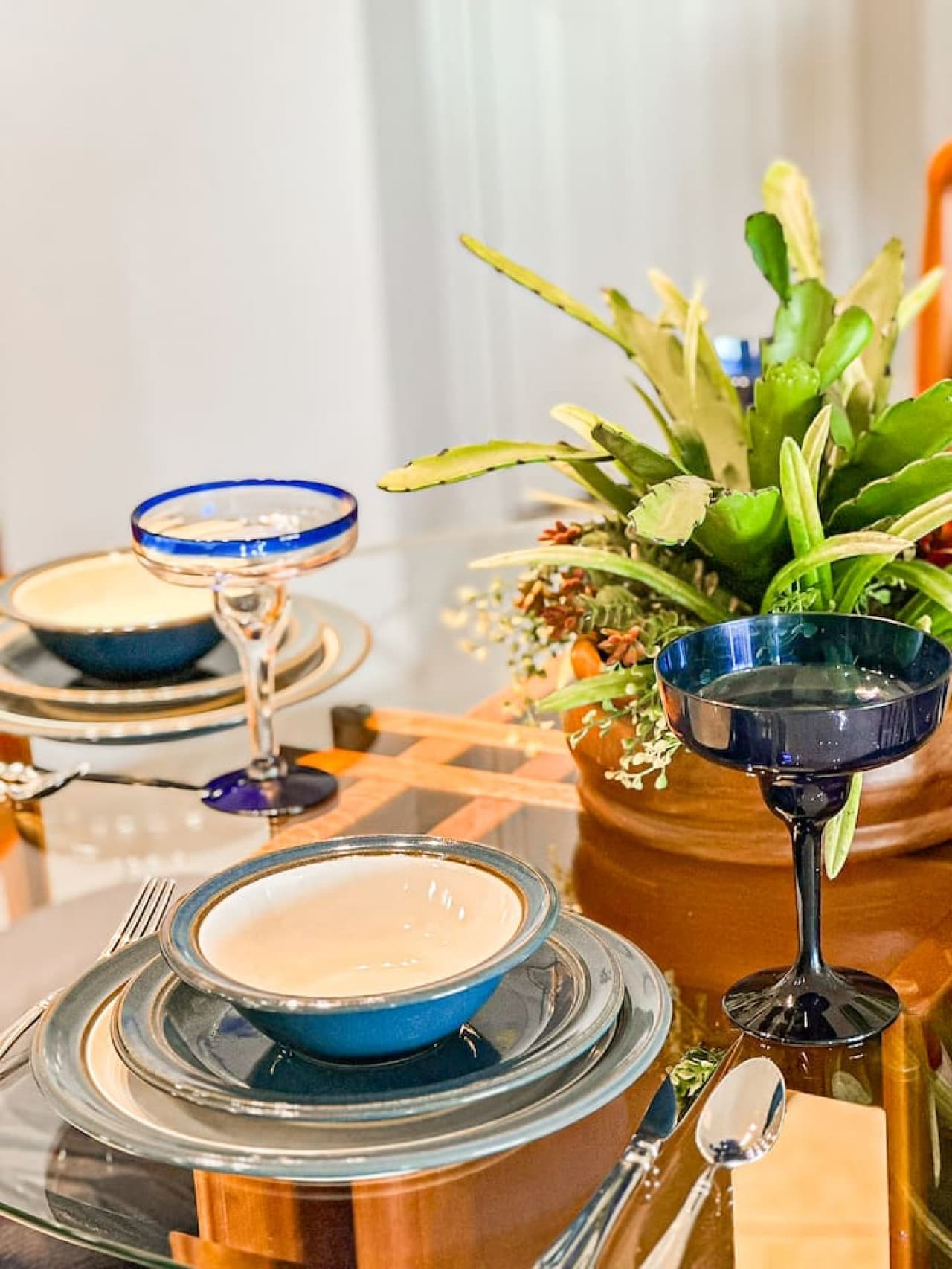 A tastefully set glass dining table with stylish dinnerware, blue-rimmed margarita glasses, and a lush greenery centerpiece, creating a charming and inviting space for meals.
