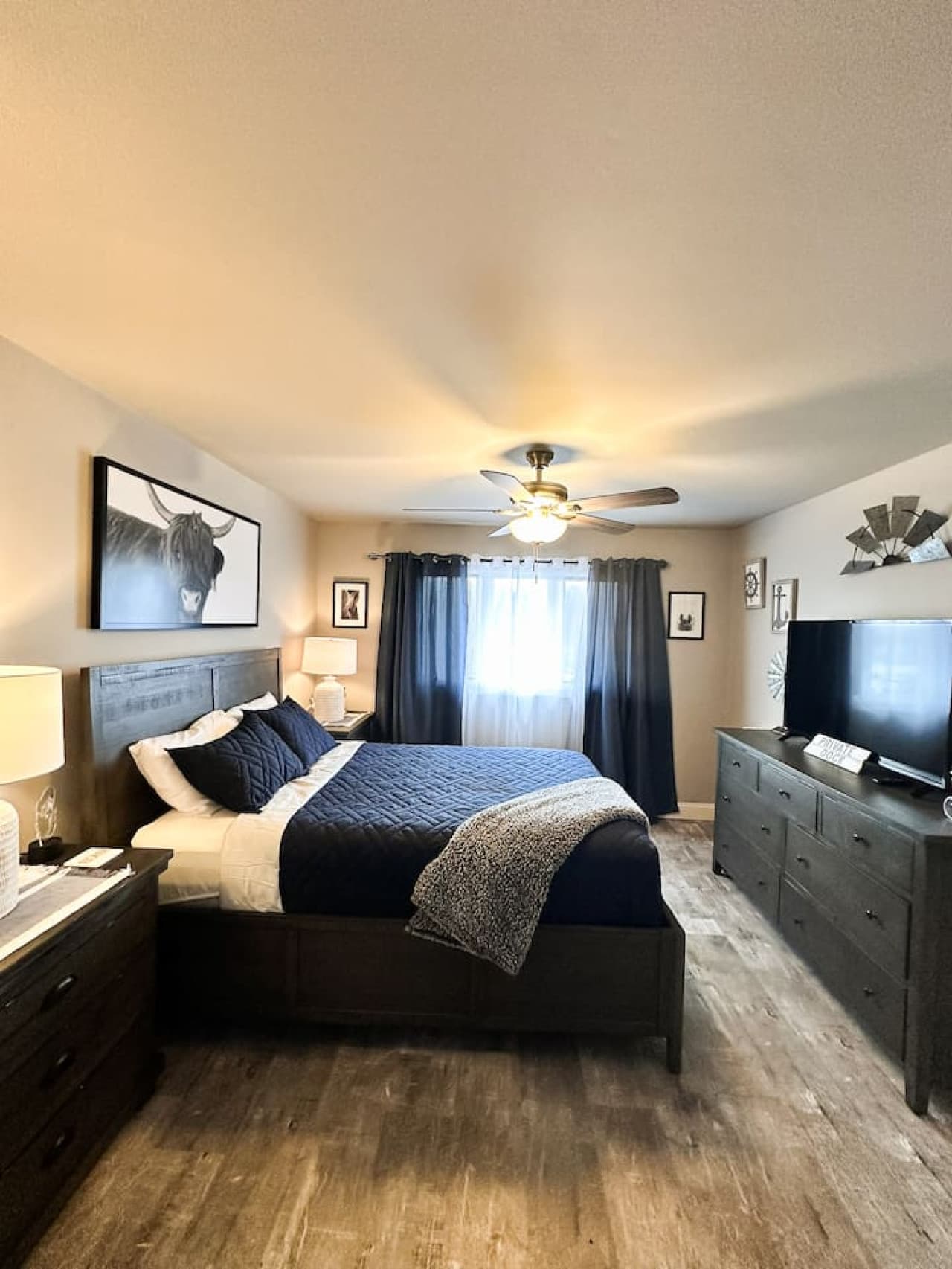 This bedroom features modern rustic charm with dark wood furnishings, a plush navy bedspread, and soft natural light filtering through blackout curtains—perfect for rest and relaxation.