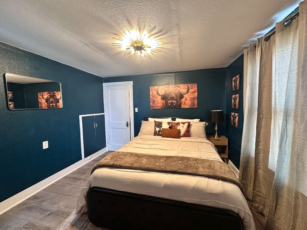 This bedroom features bold teal walls, complemented by a comfortable bed with a light grey comforter and a decorative brown throw. A large bull artwork above the bed adds character, while a mirror and a bedside table with a lamp enhance the room's fu