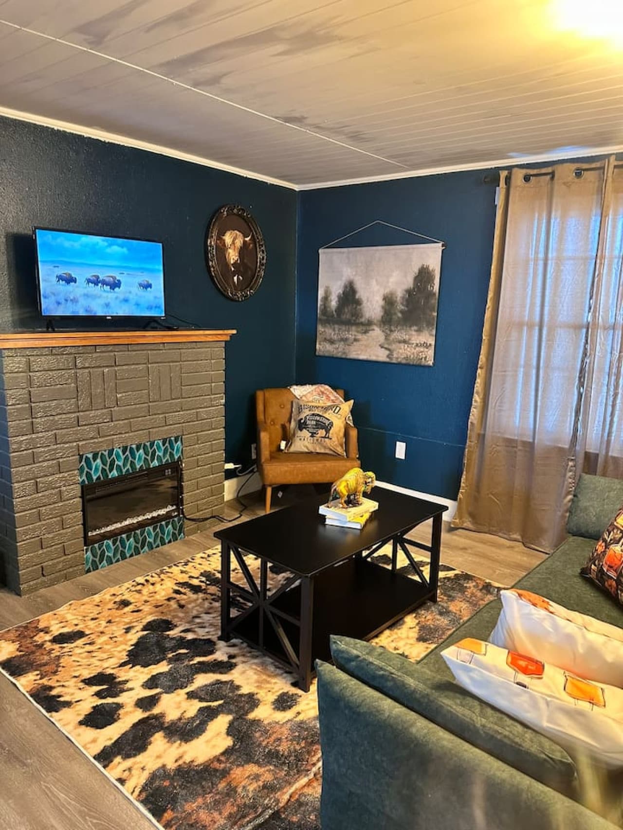This cozy living room features deep teal walls adorned with Western-themed decor, including a mounted steer skull and bison artwork above a brick fireplace with teal accents. A comfortable green sectional and a cowhide-patterned rug create a warm and