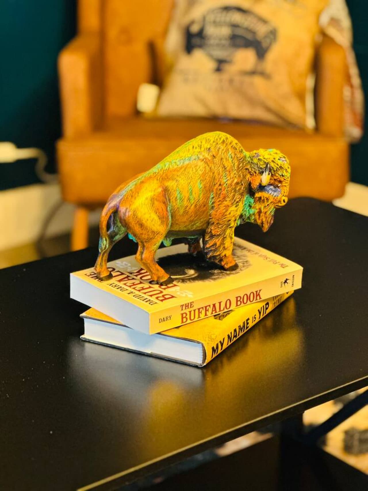 This close-up features a vibrant, colorful resin American bison figurine perched atop two books on a sleek black table, adding a touch of artistic flair and Western charm to the decor. In the blurred background, a pillow with a bison design on a tan