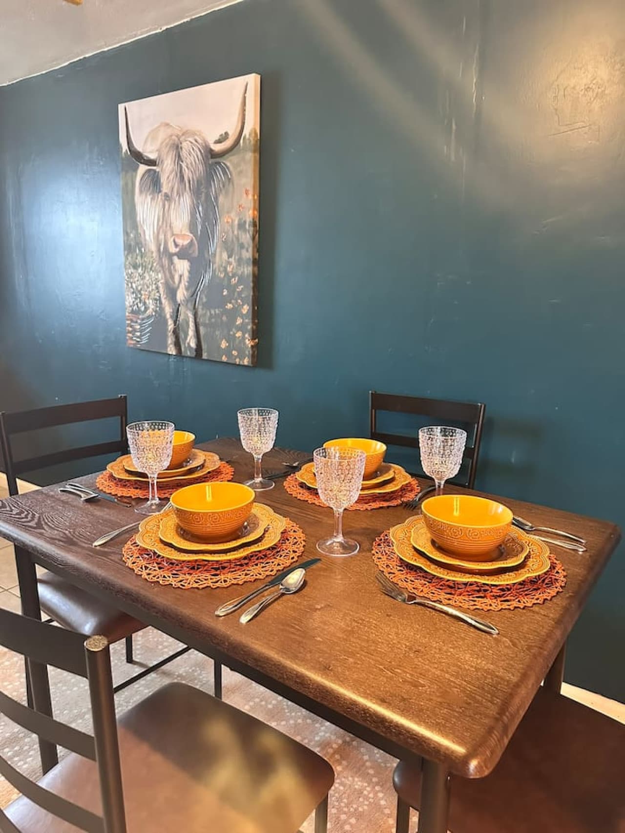 This dining area features a dark wood table elegantly set for four with orange accents against a backdrop of bold teal walls, enhanced by a striking Highland cow artwork. The setup creates a stylish and inviting space for memorable meals.