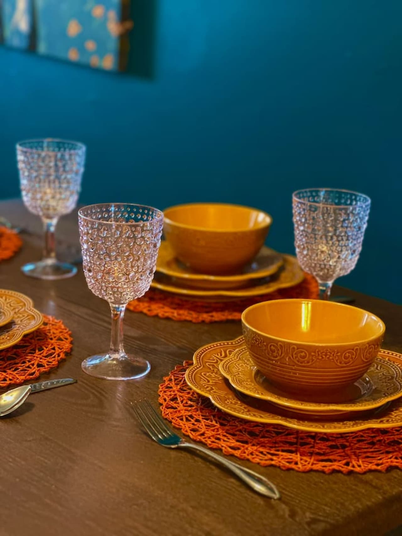 This close-up showcases a dark wood table elegantly set for four with orange accents, including intricately designed plates, bowls, and textured glassware. The vibrant orange placemats against the deep teal wall create a warm and inviting setting for