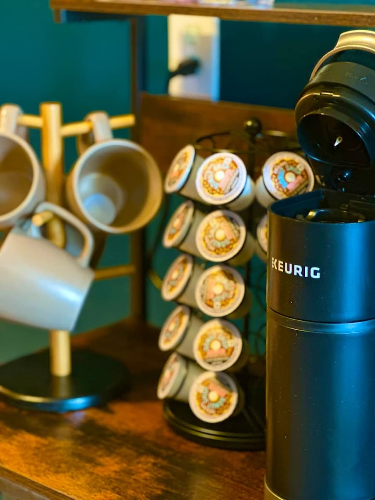 This coffee station offers convenience with a Keurig single-serve coffee maker and a carousel filled with K-Cup pods, ensuring a quick and easy start to your mornings. Mugs hang neatly on a wooden tree stand nearby, completing this functional and acc