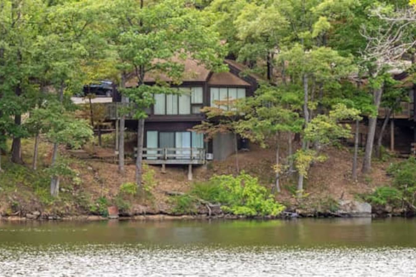 This aerial view captures a charming lake house nestled among lush trees, offering a tranquil escape. A red and white boat cruises on the water, suggesting the lake's recreational opportunities for potential renters.