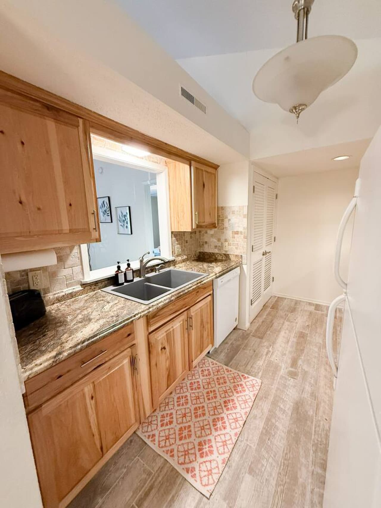 This functional kitchen features warm wood cabinetry and speckled countertops, complemented by a stylish tiled backsplash and dual sinks. The pass-through window enhances the open feel, while the durable wood-look flooring and a convenient pantry off