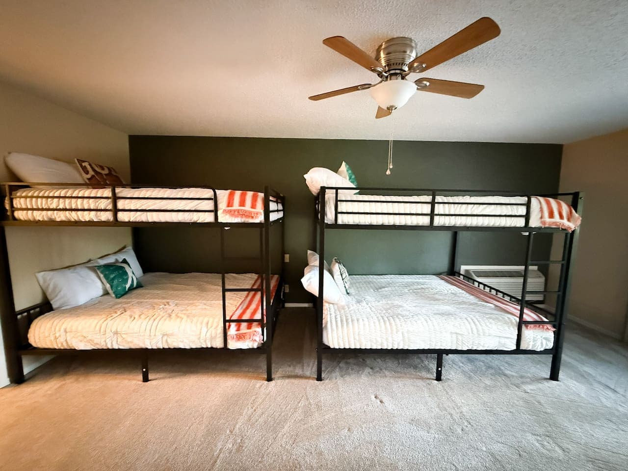 This spacious room features two sturdy black metal bunk beds, offering ample sleeping arrangements perfect for guests or a large family. A neutral carpet and a striking dark green accent wall complement the functional design, while a ceiling fan ensu