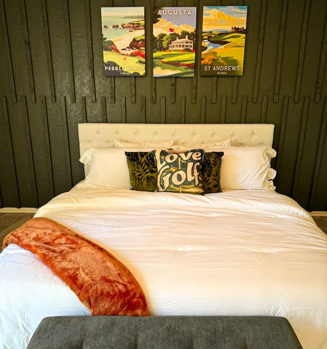 This stylish bedroom features a plush, tufted headboard and a collection of golf-themed art, perfect for enthusiasts. The room's dark accent wall provides a sophisticated backdrop, complemented by the bright white bedding and a cozy throw, creating a