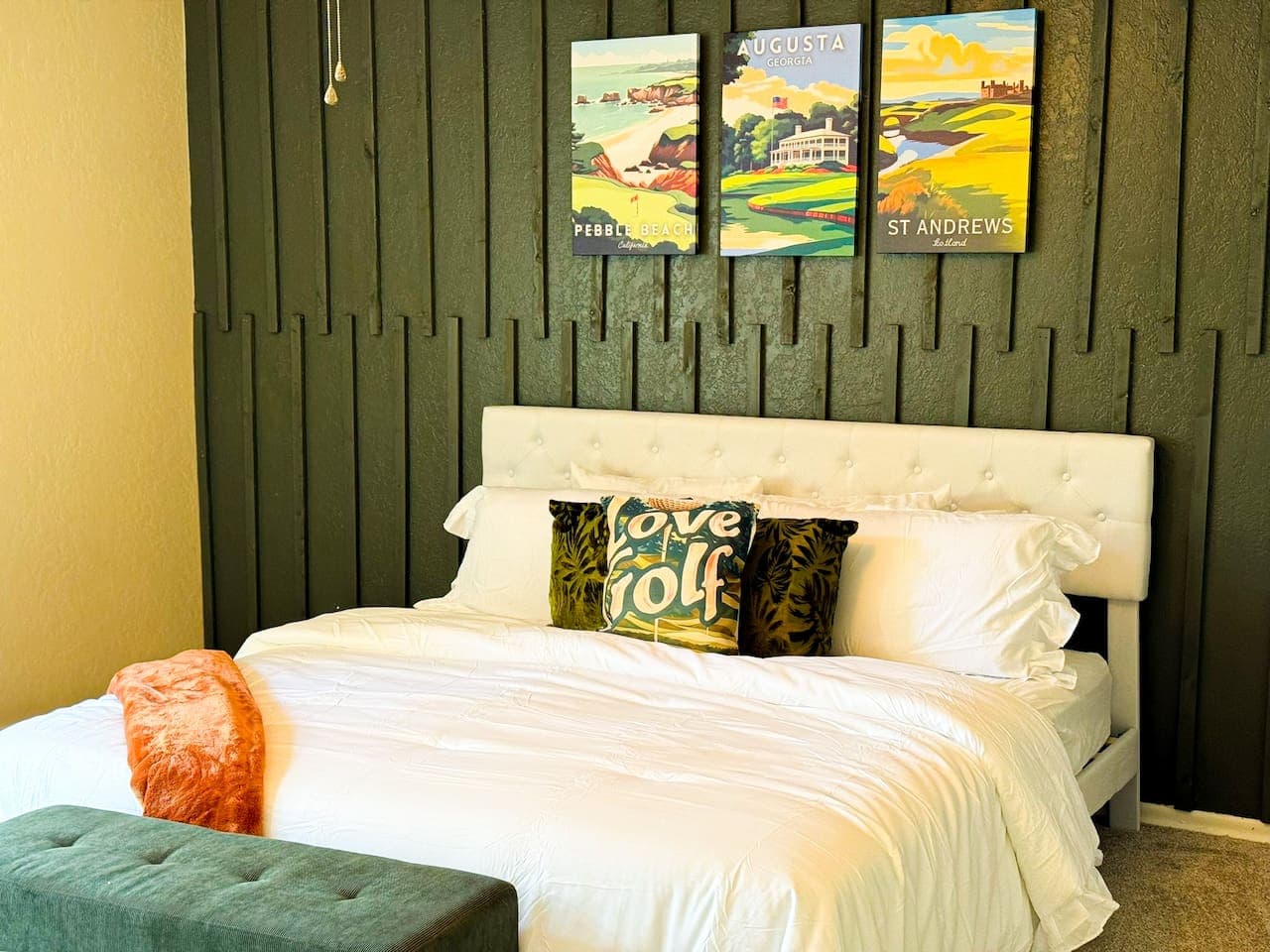 This inviting bedroom features a plush white upholstered bed, beautifully contrasted by a stylish olive green board and batten accent wall. The space is thoughtfully decorated with golf-themed artwork, adding a unique and sophisticated touch.