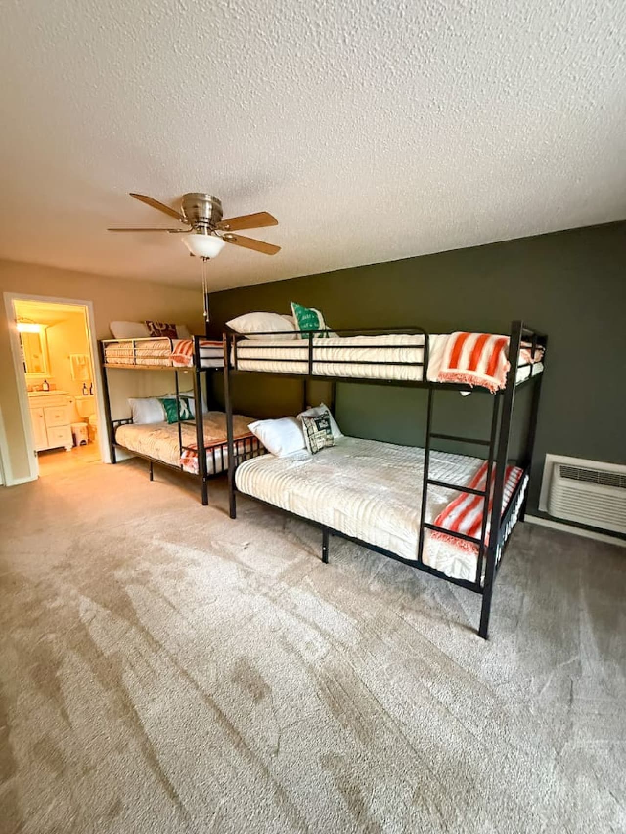 This spacious room features two sturdy black metal bunk beds, offering ample sleeping arrangements perfect for guests or a large family. A neutral carpet and a striking dark green accent wall complement the functional design, while a ceiling fan ensu