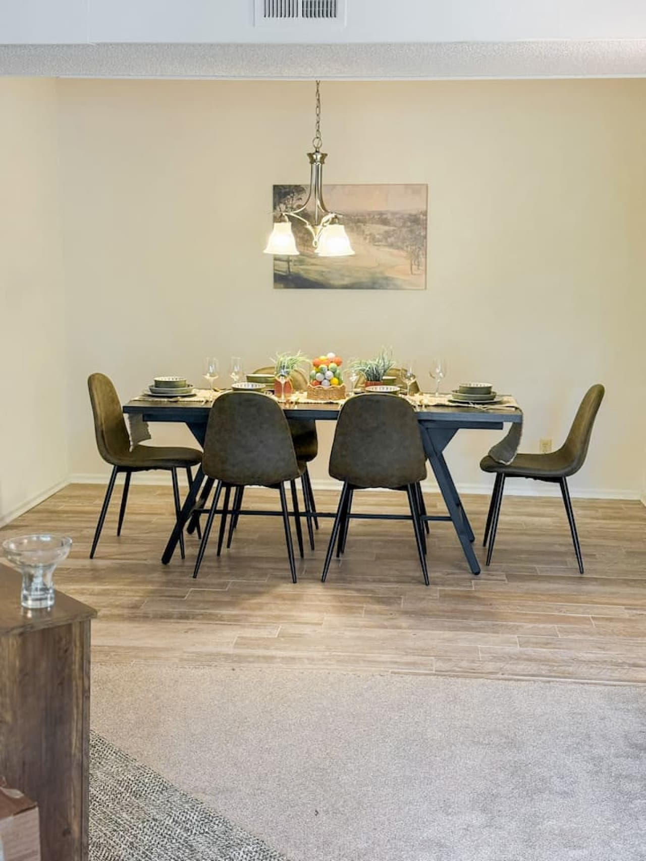 This elegant dining area features a modern table surrounded by stylish olive green chairs, perfectly illuminated by a classic chandelier. The space seamlessly transitions from durable hard flooring to a plush carpeted area, offering a versatile layou