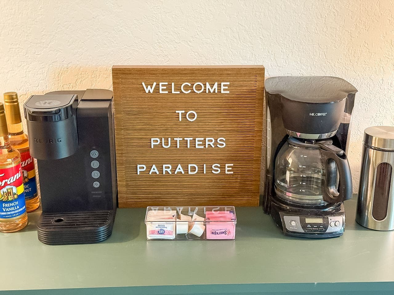 This charming coffee station welcomes guests with a sign that reads "WELCOME TO PUTTERS PARADISE," suggesting a golf-themed retreat. It features both a Keurig machine and a traditional drip coffee maker, along with various flavor syrups and sweetener
