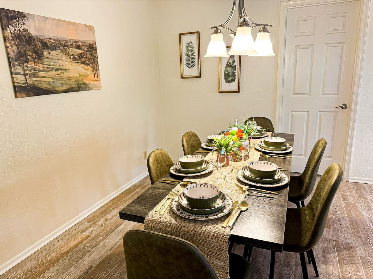This beautifully set dining room features a dark wood table with seating for six, adorned with a woven runner and elegant place settings. The golf-themed centerpiece adds a playful touch, while framed botanical prints and a large landscape painting o