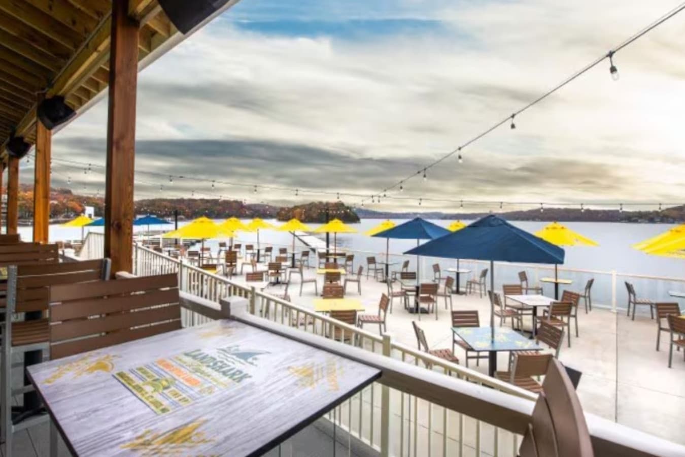 This expansive lakeside dining terrace offers breathtaking water views and abundant outdoor seating under a mix of sunny yellow and deep blue umbrellas. Perfect for a vibrant restaurant or event venue, the space provides a quintessential waterfront e