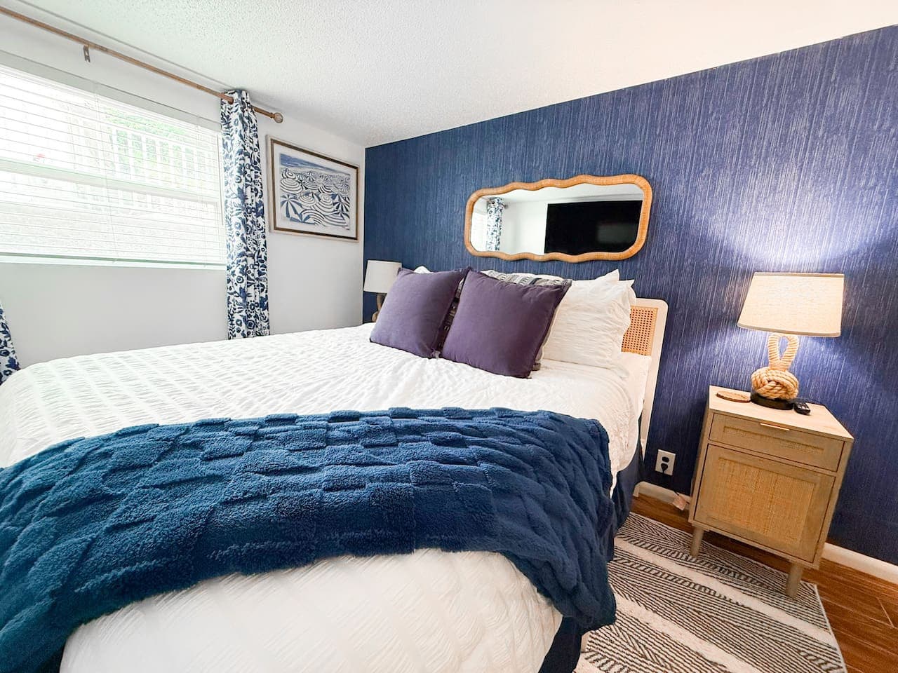 A spacious queen-size bed sits against a backdrop of deep blue wallpaper, evoking a sense of calm and sophistication. A huge window floods the room with natural light, a perfect balance of coziness and openness.