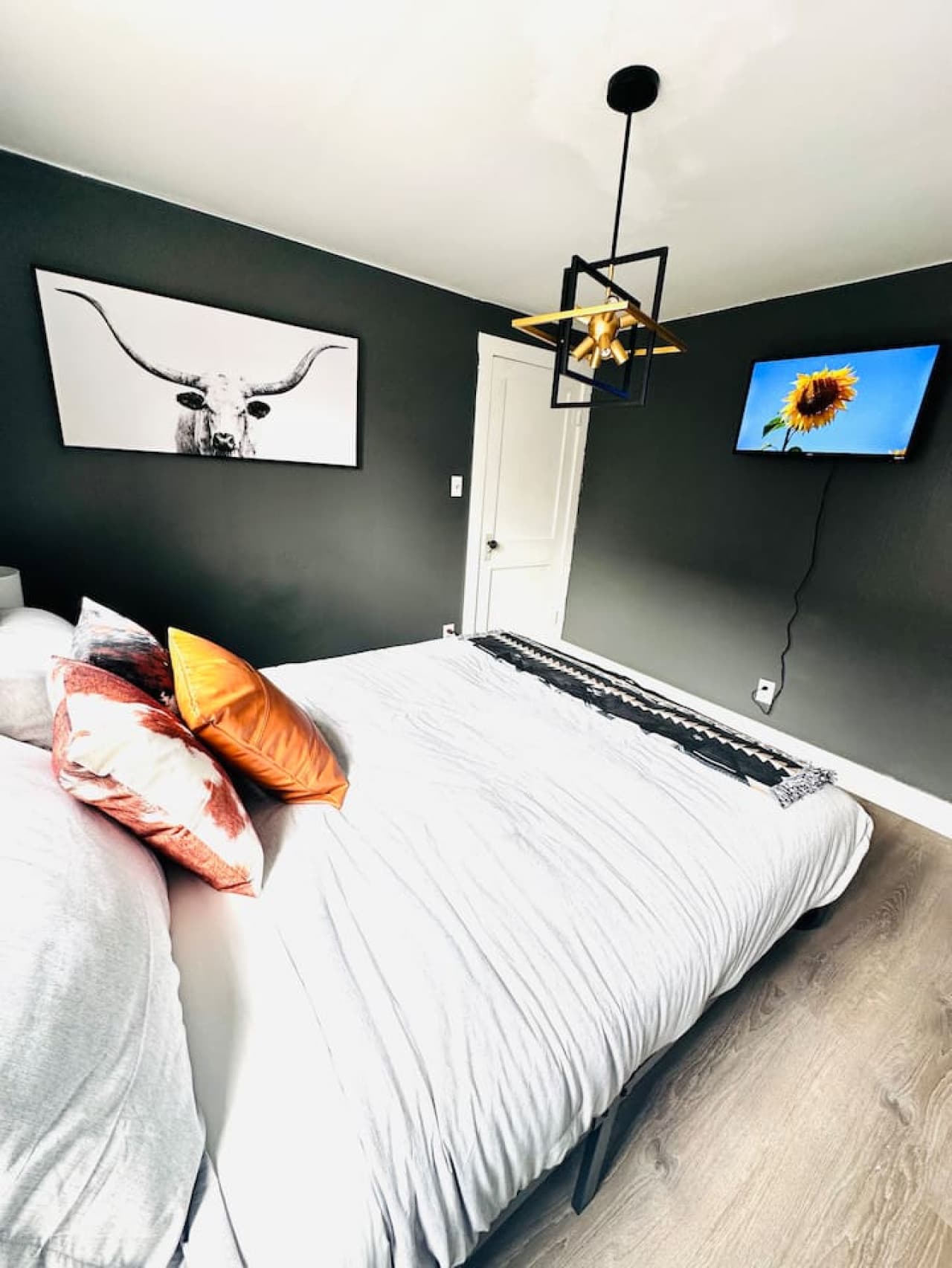 This spacious bedroom features a comfortable bed with modern bedding, accented by stylish decorative pillows. The room is thoughtfully decorated with unique wall art, including a striking longhorn print and a vibrant sunflower on the mounted TV, crea