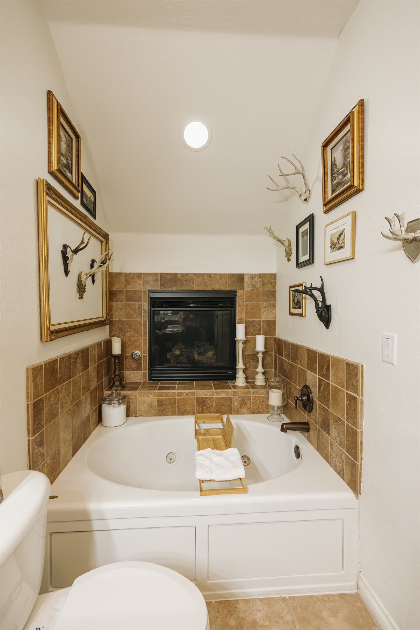 Soak beneath a glowing fireplace in this spa-like bathroom retreat. The deep jetted tub is framed by warm stone tile, curated vintage accents, plush towels, and soft lighting—creating a serene, romantic space to unwind after a day in the White Mountains.